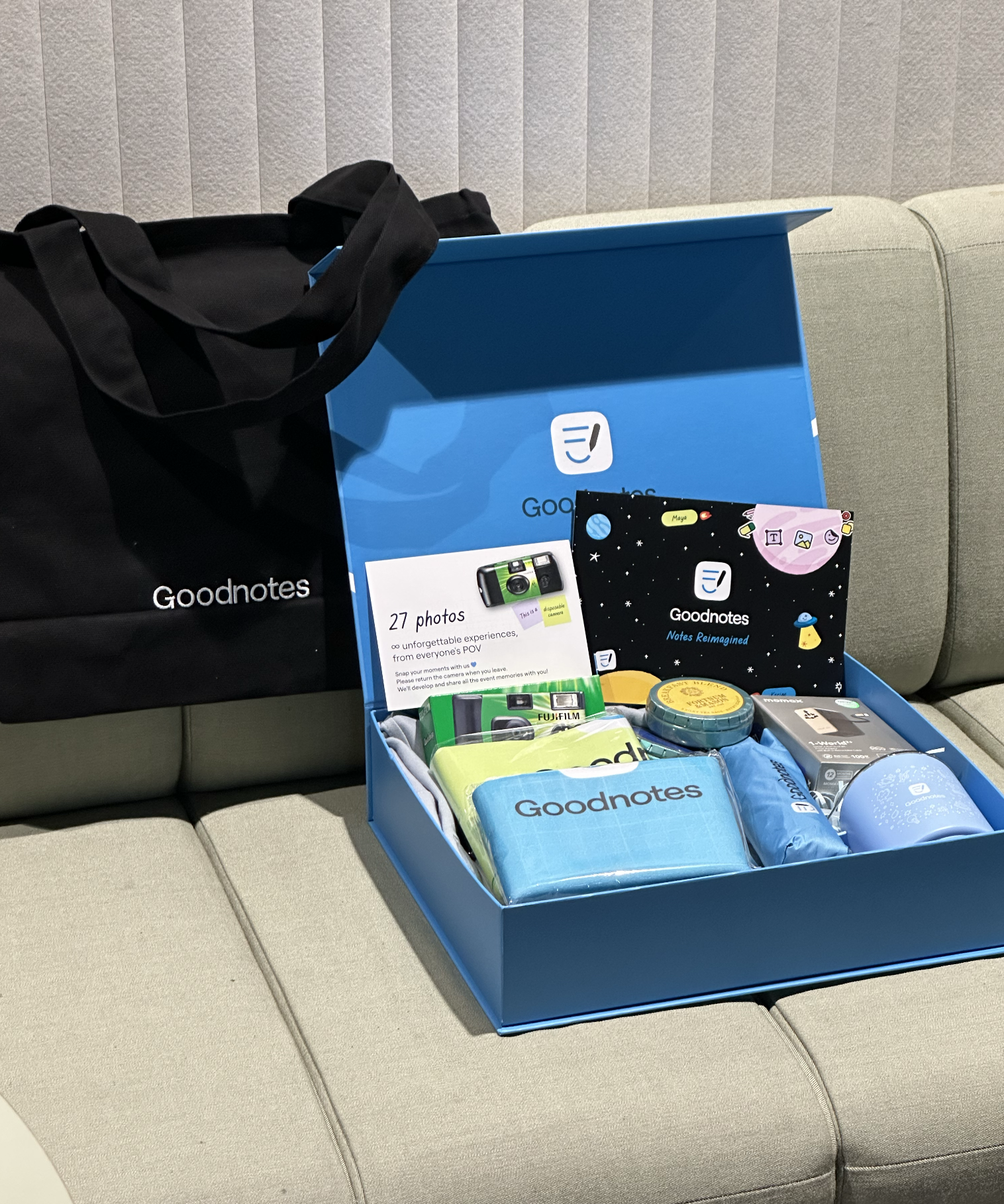 An open merch kit: disposable camera, notebook, umbrella, tumbler, tea and travel adapter, beside the Goodnotes tote