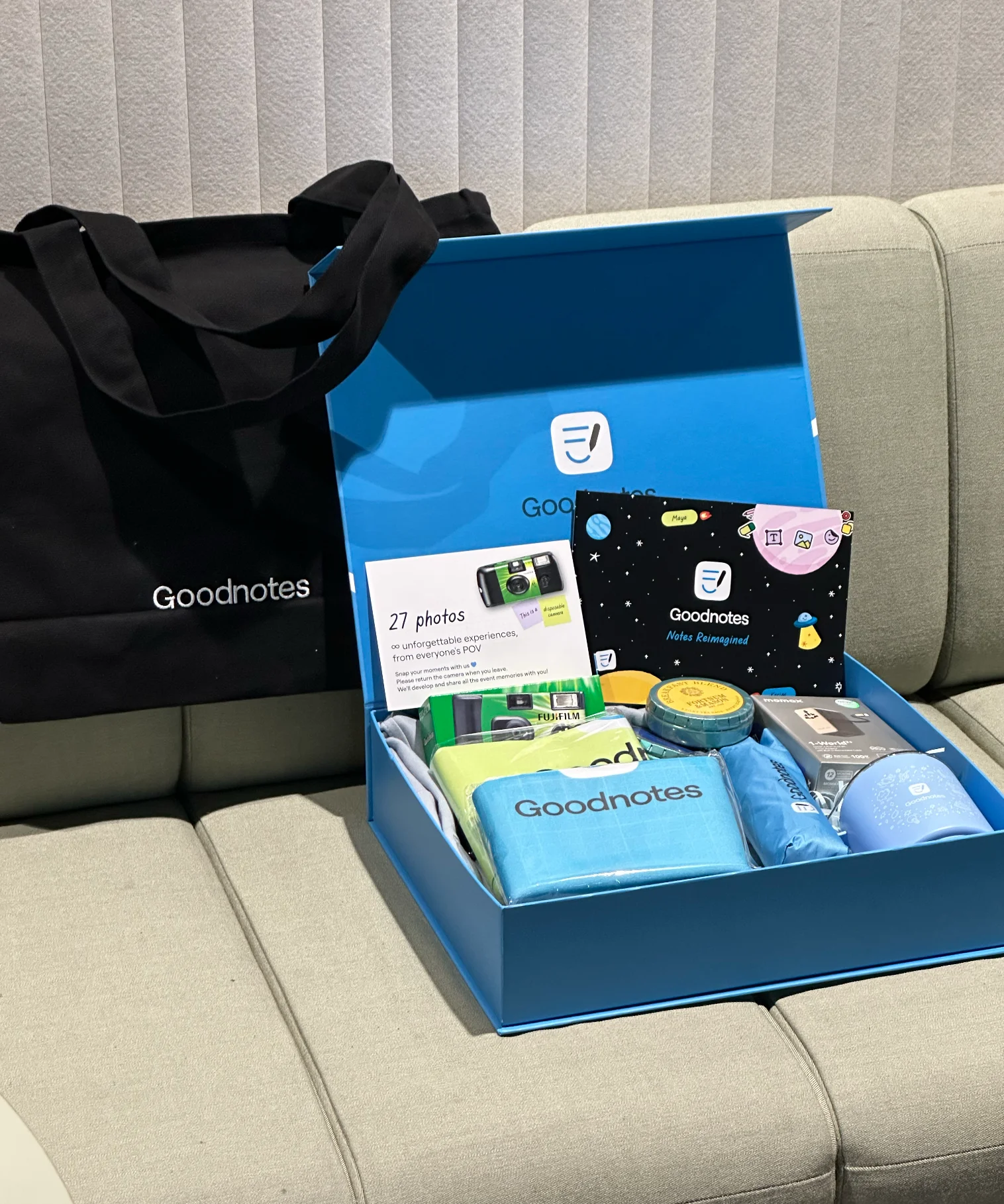 An open merch kit: disposable camera, notebook, umbrella, tumbler, tea and travel adapter, beside the Goodnotes tote